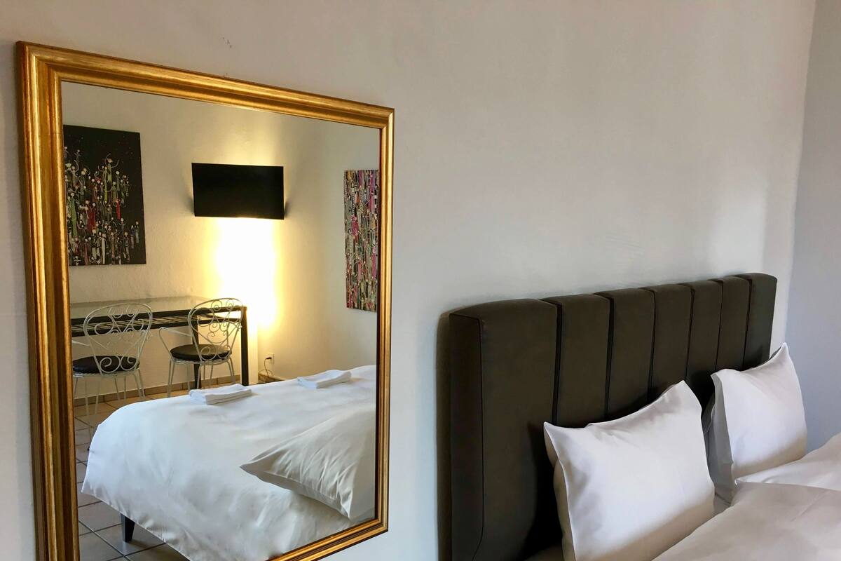 A hotel room with a large mirror, a bed with white linens, a headboard, and two chairs.