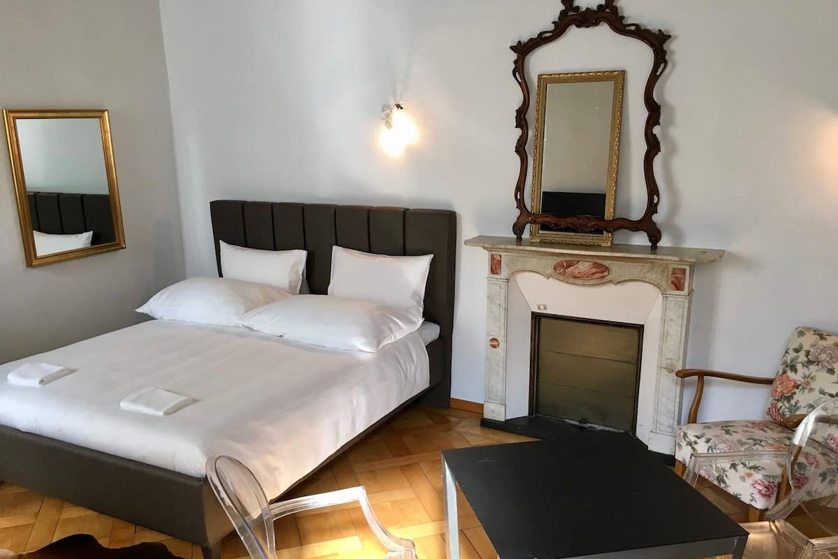 A neatly made bed with white linens, a wooden headboard, and two bedside tables. A clear plastic chair and a black table are placed in front of a fireplace with a marble mantle.