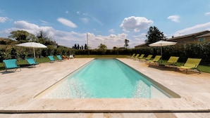 Una piscina rectangular con agua cristalina, rodeada de sillas de descanso y sombrillas, con árboles y un cielo despejado de fondo.