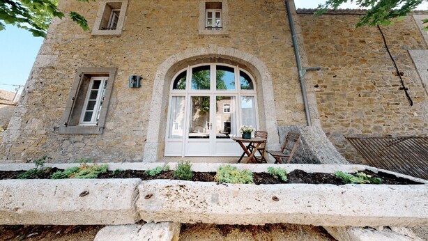 Un edificio de piedra con un amplio ventanal de arco, una pequeña mesa con sillas y un jardín delantero.