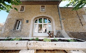 Un edificio in pietra con un'ampia finestra ad arco, un piccolo tavolo con sedie e un'area giardino di fronte.