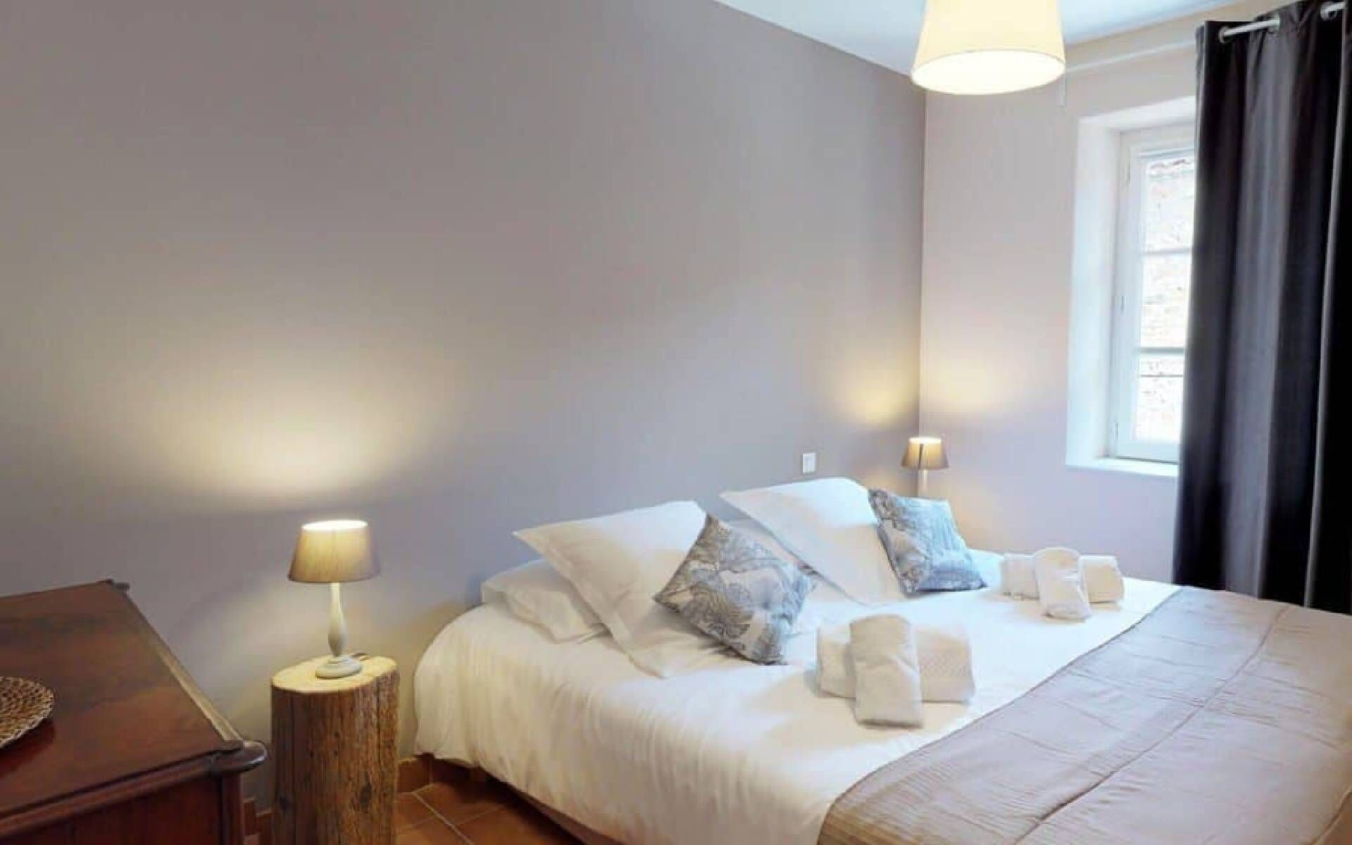 A bedroom with a bed, bedside lamps, a wooden nightstand, and a window with curtains.