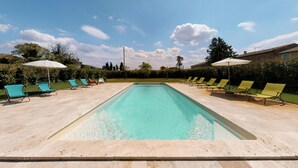 Una piscina rectangular con agua cristalina, rodeada de sillas de descanso y sombrillas, con árboles y un cielo despejado de fondo.