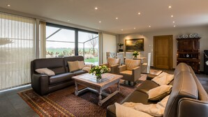 A spacious living room with a large window, a brown leather sofa, a coffee table, and a wooden cabinet. - Polderwoning Cleylantshof max. 8 p midden in de polders van het Meetjesland. (Watervliet)