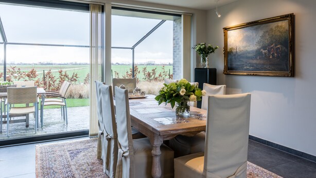 A dining area with a table set for four, chairs, a vase with flowers, and a painting on the wall. - Polderwoning Cleylantshof max. 8 p midden in de polders van het Meetjesland. (Watervliet)
