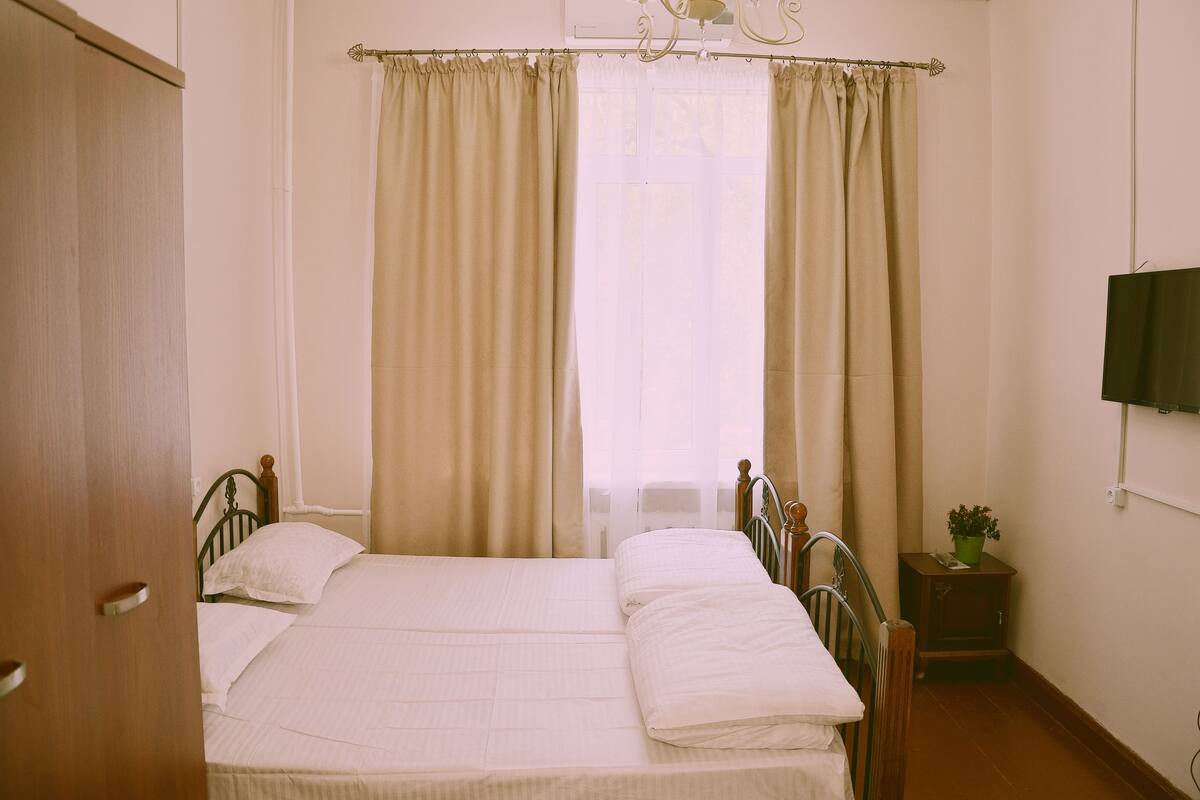 A bedroom with a bed, curtains, a wooden wardrobe, a small bedside table with a plant, and a wall-mounted TV.