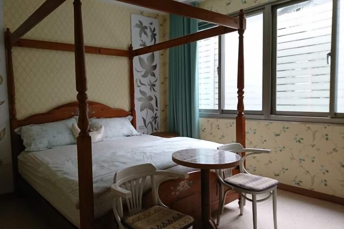 A four-poster bed with a wooden headboard, a small round table with two chairs, and a window with blinds.