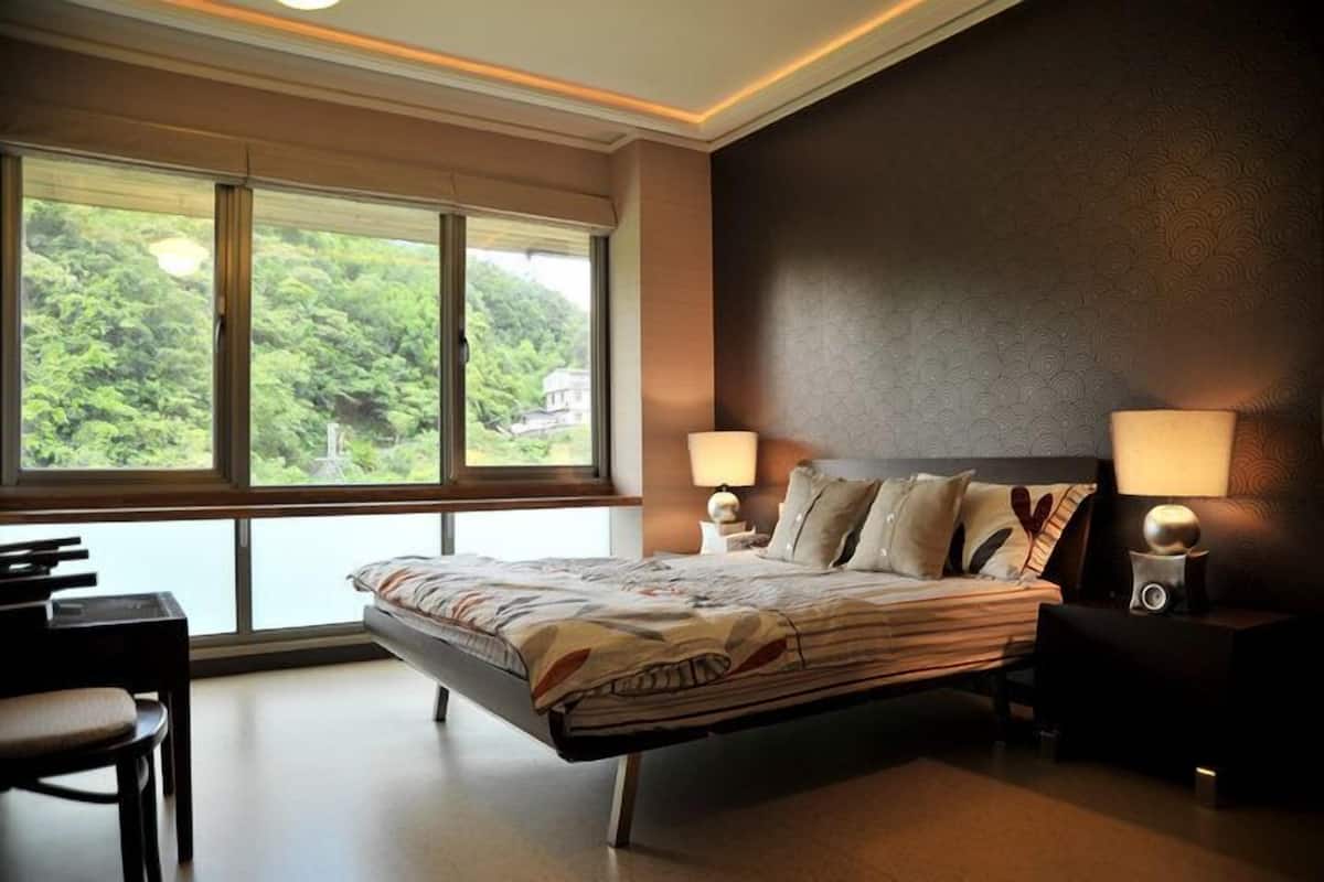 A modern hotel room with a large bed, two bedside lamps, a desk, and a view of lush greenery through large windows.