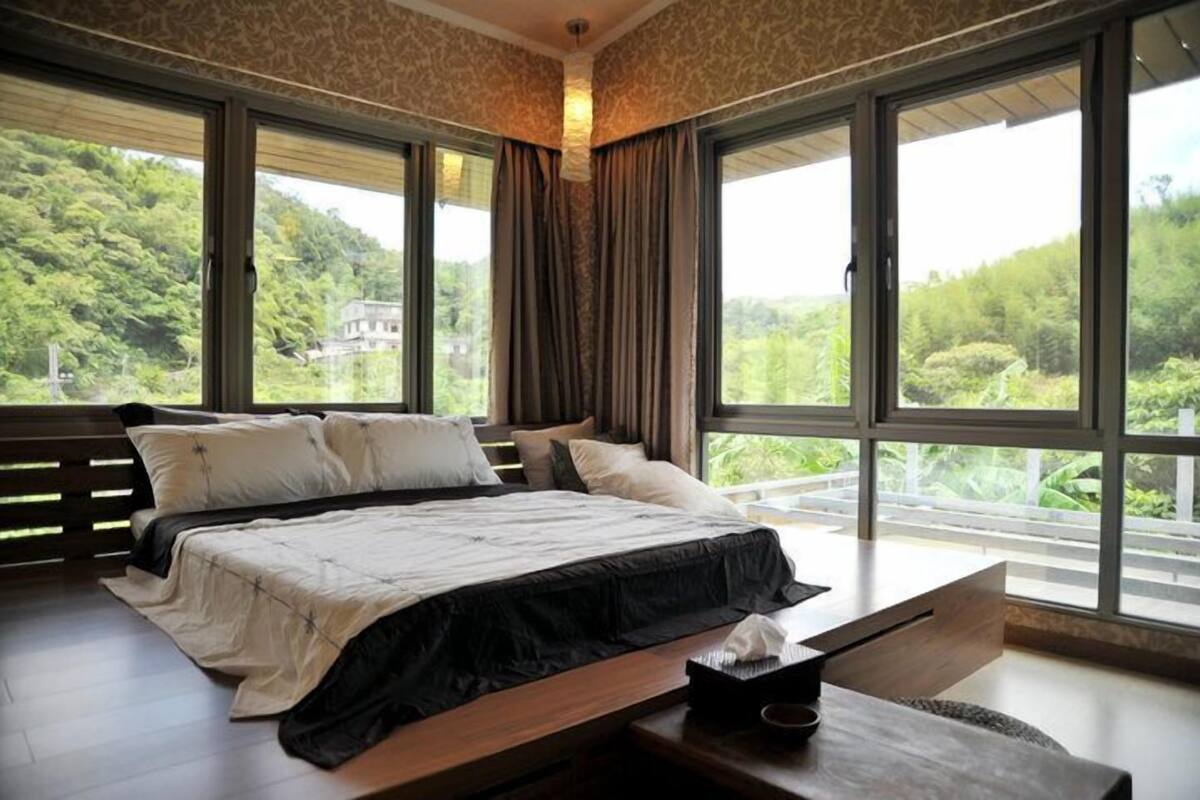 A bedroom with a large bed, a wooden bench, and a view of a lush green landscape through large windows.