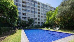 A swimming pool surrounded by a grassy area with trees and a multi-story building in the background. - Central Urban Oasis With Garden View & Swimming Pool (Cape Town)