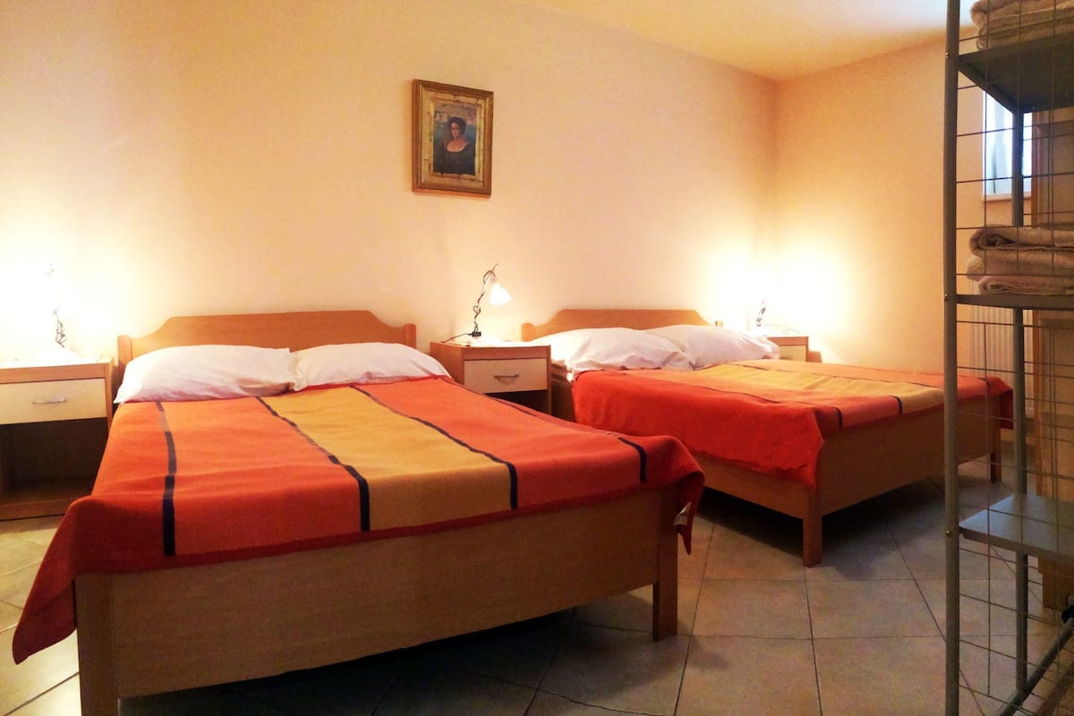 A hotel room with two beds, each with a wooden headboard and a red and beige bedspread. There is a nightstand with a lamp and a framed picture on the wall.