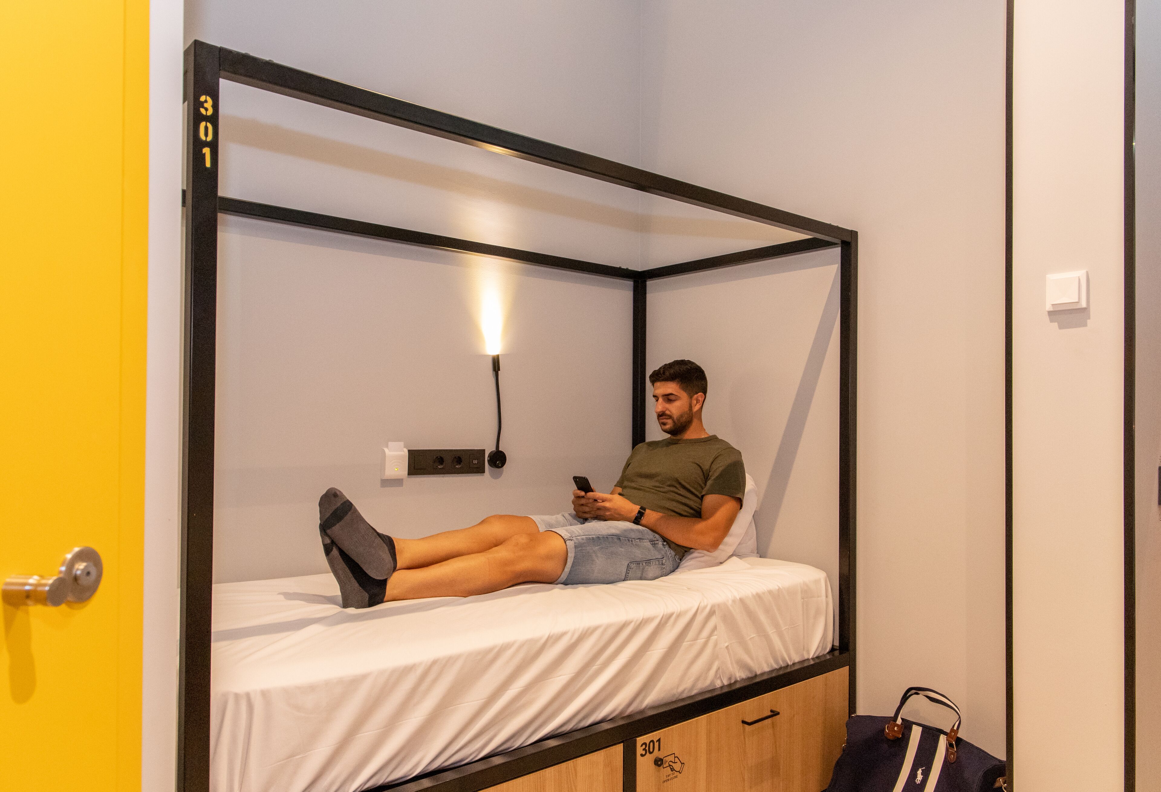 A single bed in a room with a yellow door, a wall-mounted light, and a small cabinet with the number 301.