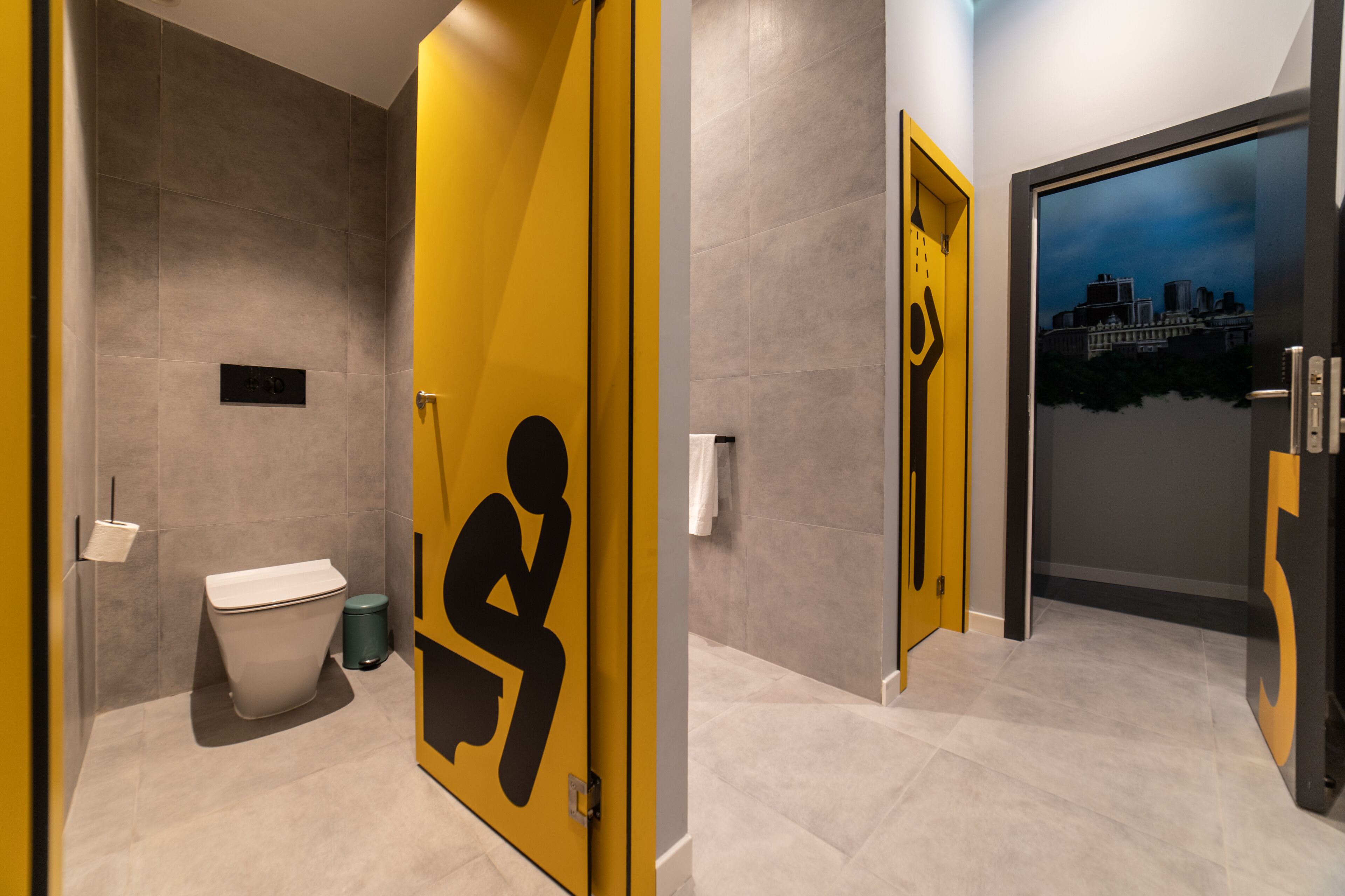 Un baño moderno con inodoro, un cesto de basura y una puerta amarilla con el símbolo de figura sentada.