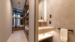 Una habitación de hotel moderna con literas, suelo de madera, un ventanal grande y un baño con lavabo y espejo.