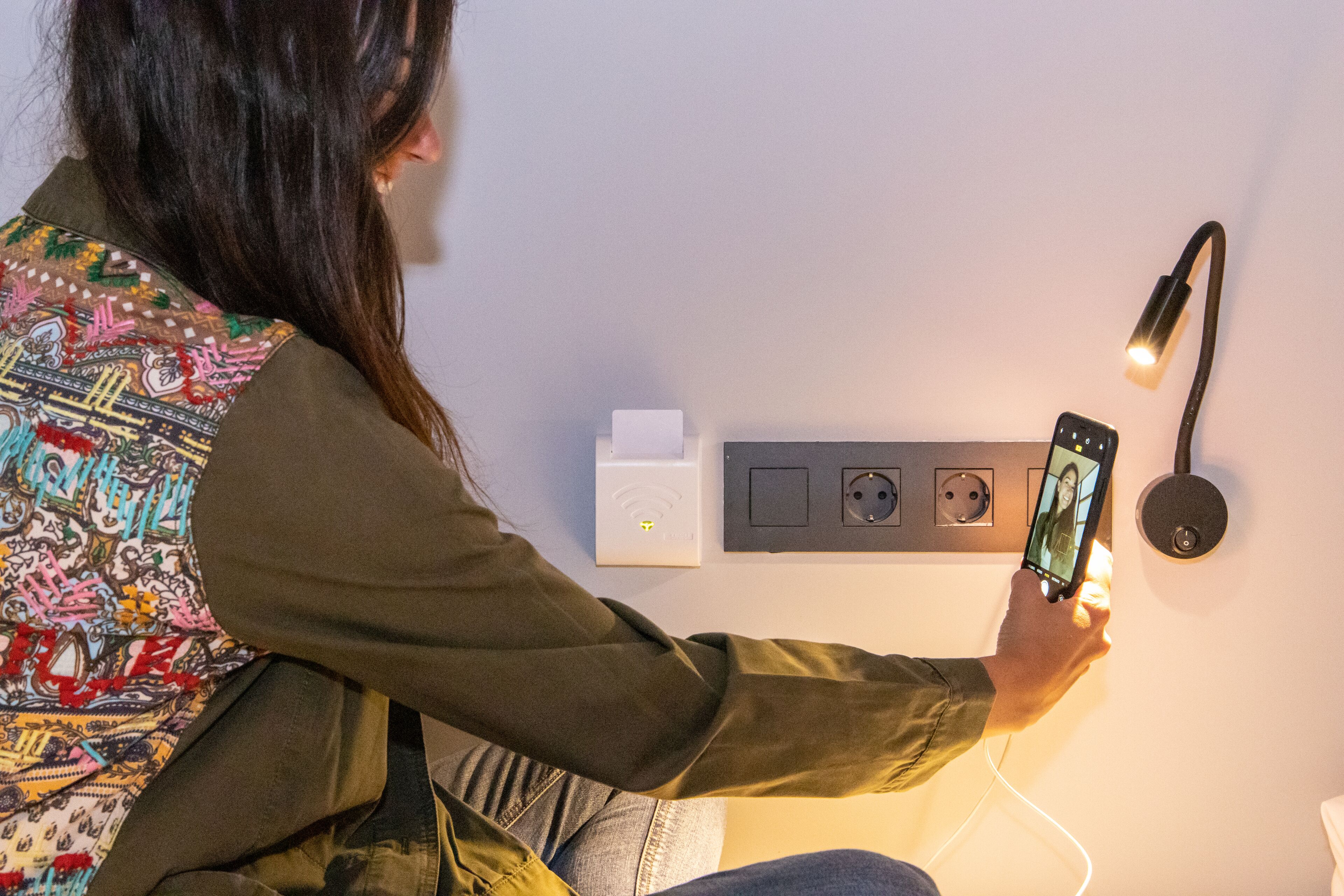 Une personne utilise un smartphone dans une pièce où se trouvent une station de recharge fixée au mur et une lampe.