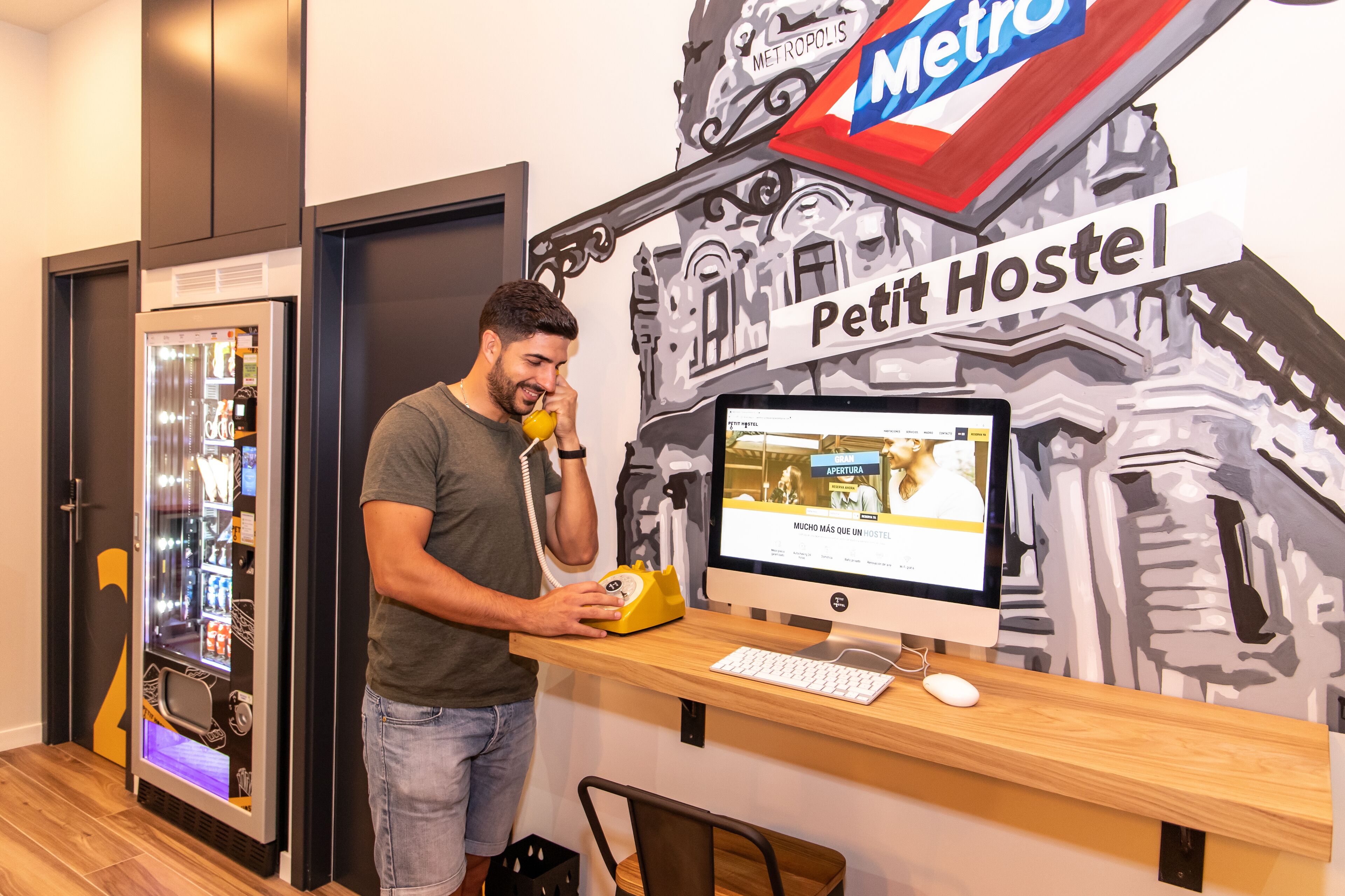 A man at a desk with a computer, a vending machine, and a wall with a 'Petit Hostel' sign.