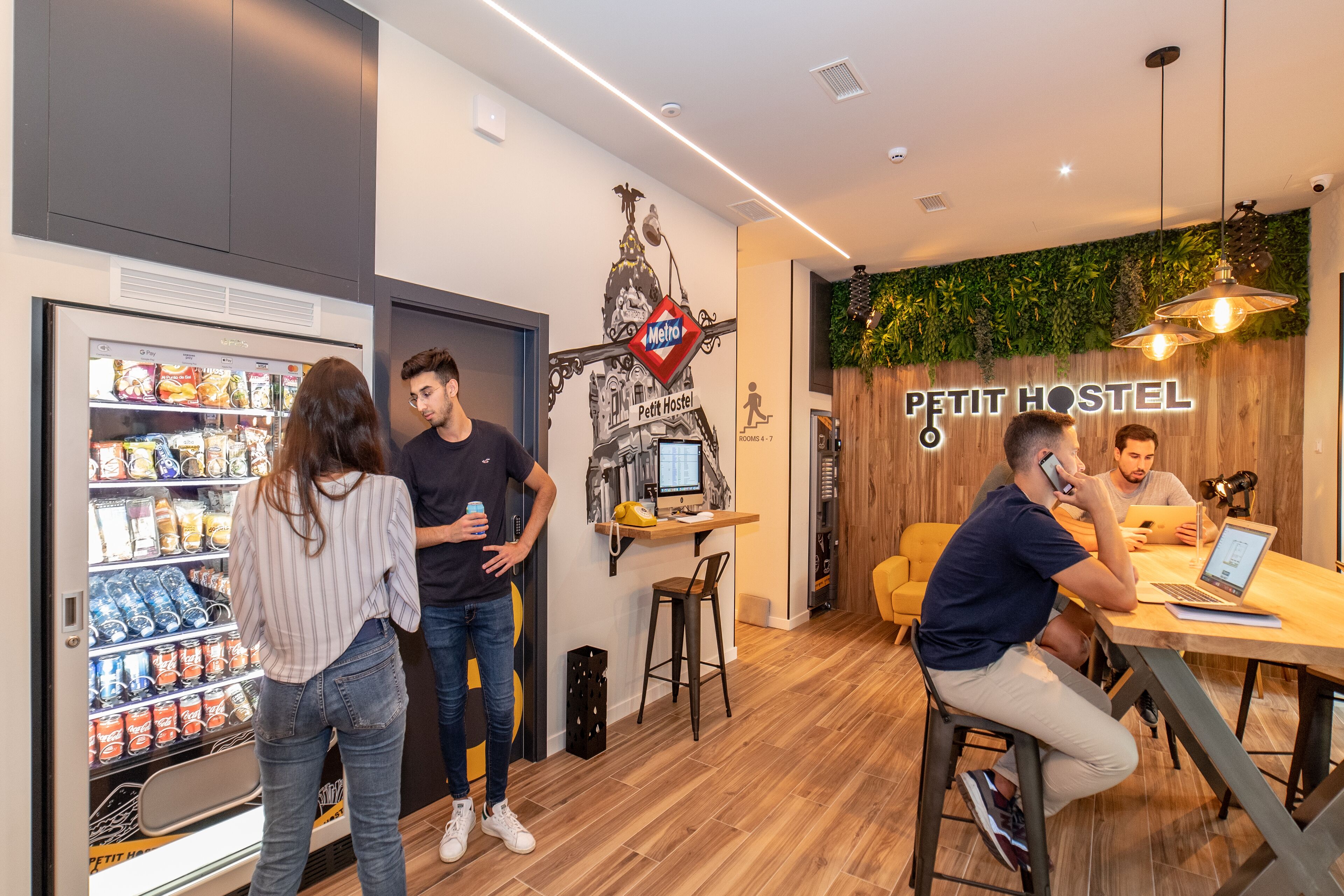 A modern interior with a vending machine, a wooden wall with a 'Petit Hostel' sign, and a seating area with a laptop.