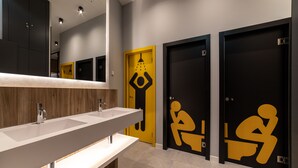 Un baño moderno con un espejo grande, dos lavamanos y tres puertas con símbolos de baño.