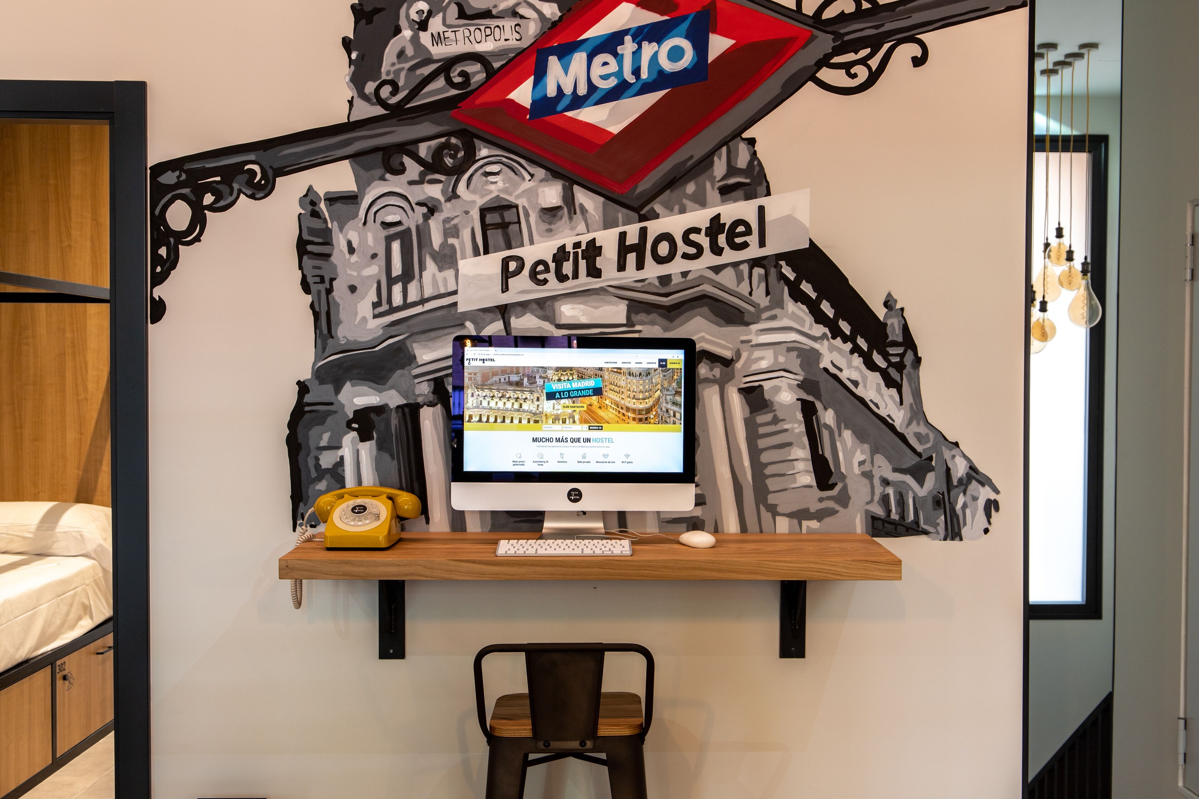 A room with a desk featuring a computer, a telephone, and a chair. A wall decoration includes a 'Metro' sign and a 'Petit Hostel' sign.
