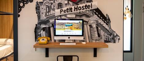 A room with a desk featuring a computer, a telephone, and a chair. A wall decoration includes a 'Metro' sign and a 'Petit Hostel' sign.