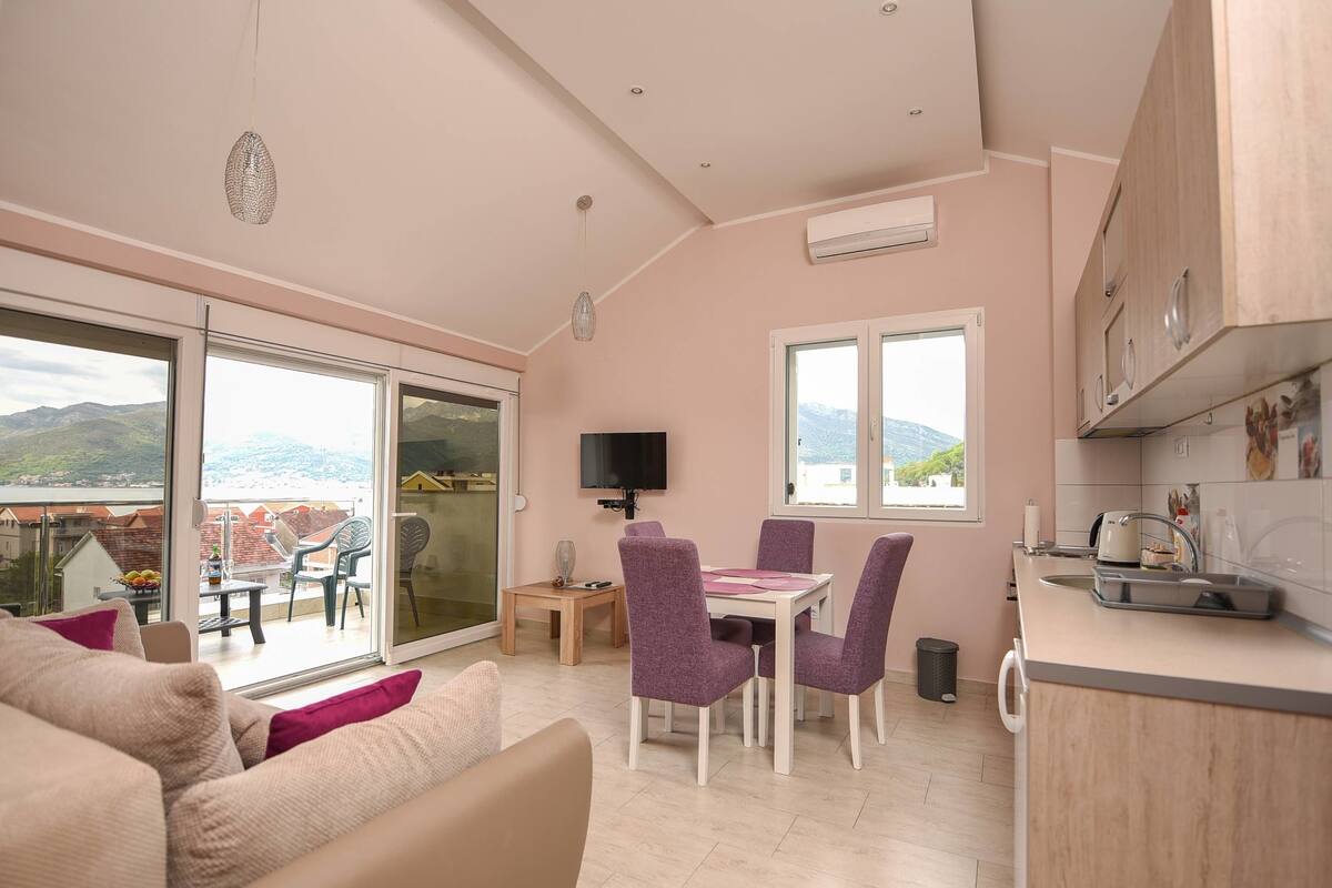 Una sala de estar moderna con un sofá, una mesa de comedor con sillas moradas, un televisor y una zona de cocina con armarios y un ventanal.