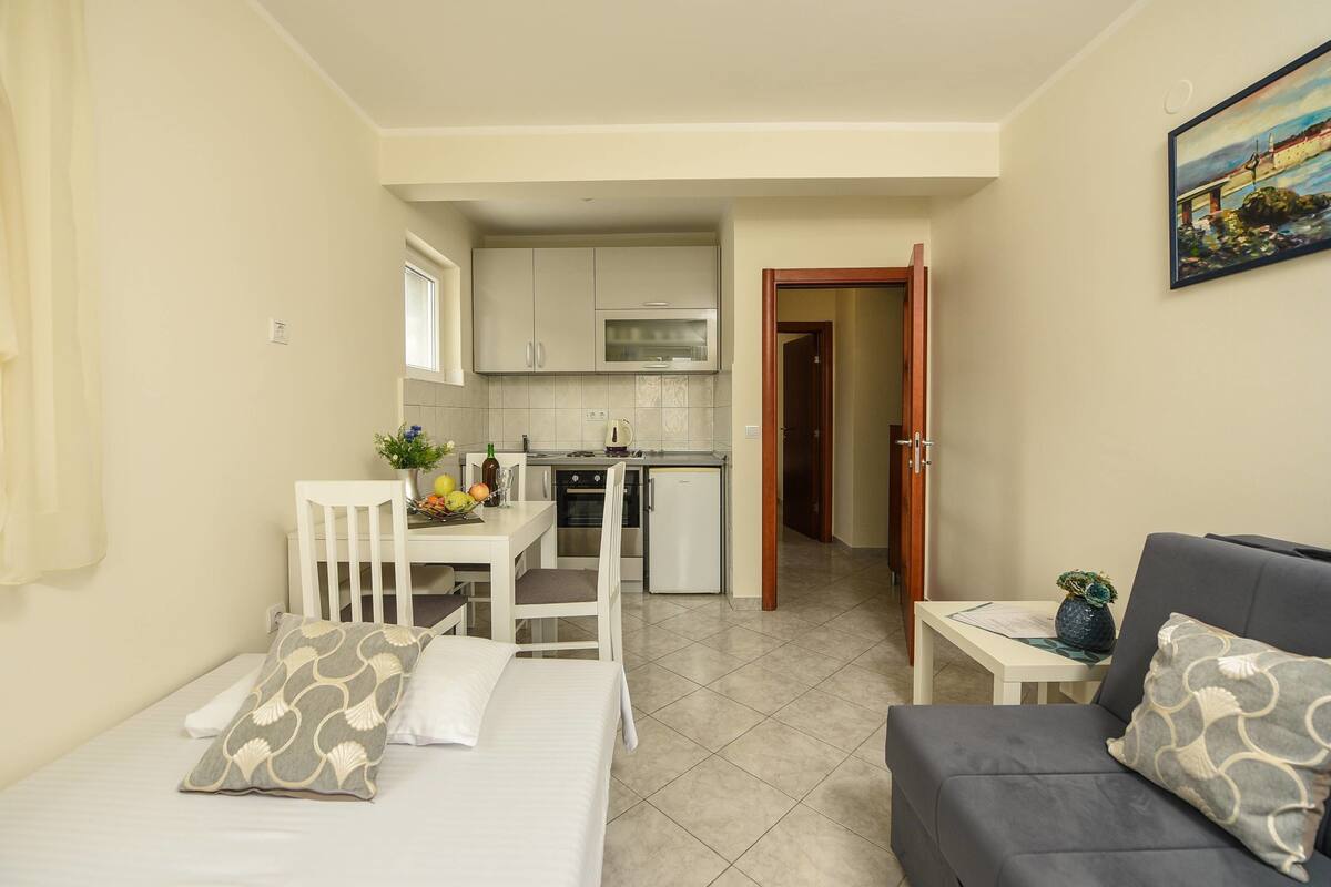 Una habitación compacta con cama, sofá, una mesita, kitchenette y una puerta que da a otra habitación.