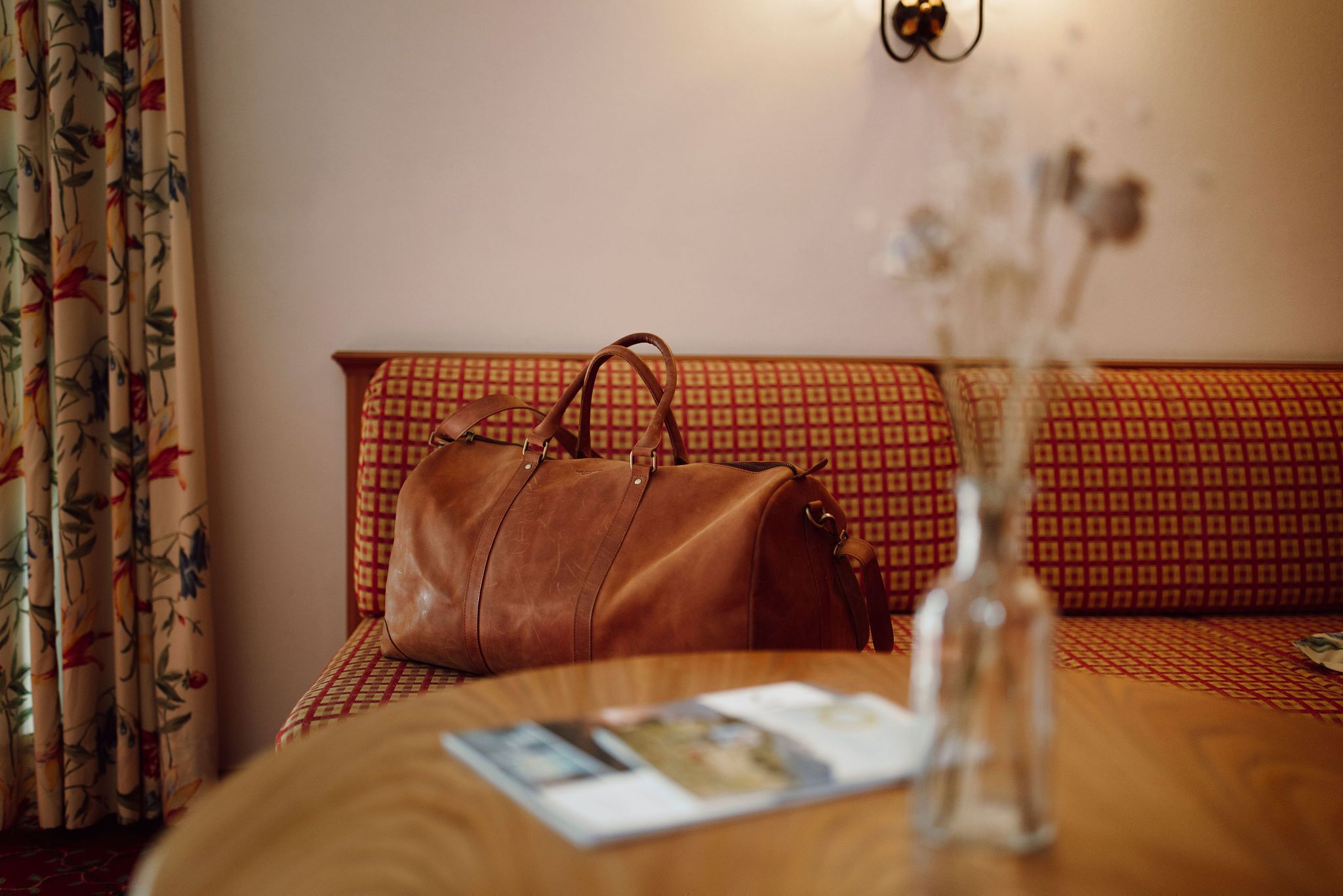 Una habitación con una mesa de madera, un bolso de cuero marrón, un jarrón con flores secas y un sofá estampado.