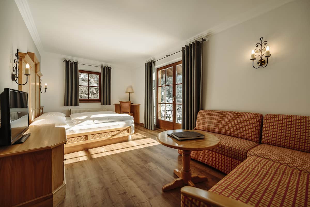 Habitación de hotel con cama, sofá, televisión, lámpara y ventana con cortinas.