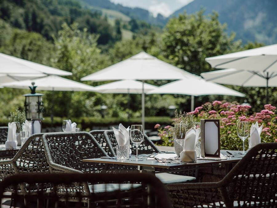 An outdoor dining area with wicker chairs and tables, white umbrellas, and a mountain backdrop.