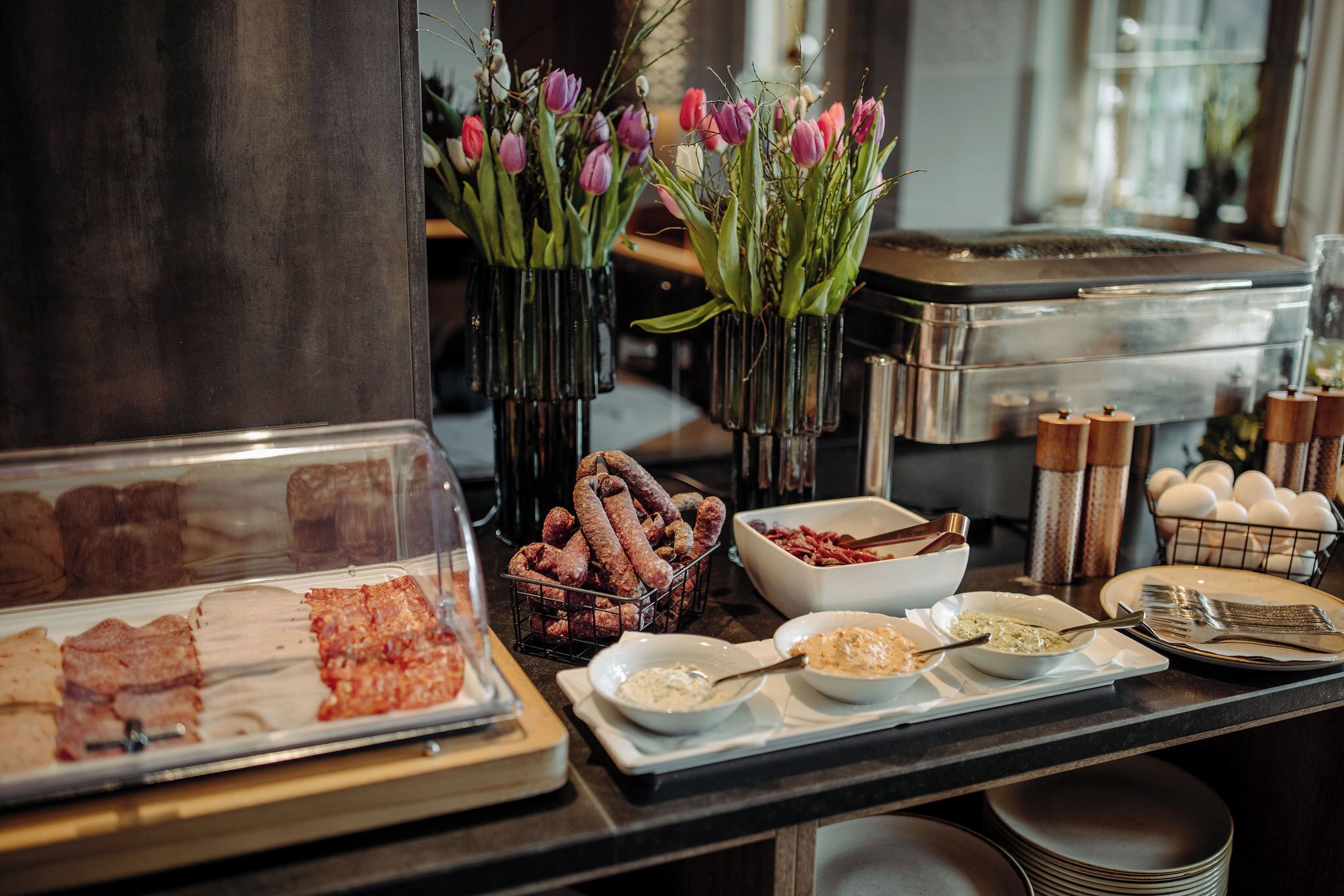 Buffet com diversos alimentos, incluindo carnes e patês, e vasos decorativos com flores.