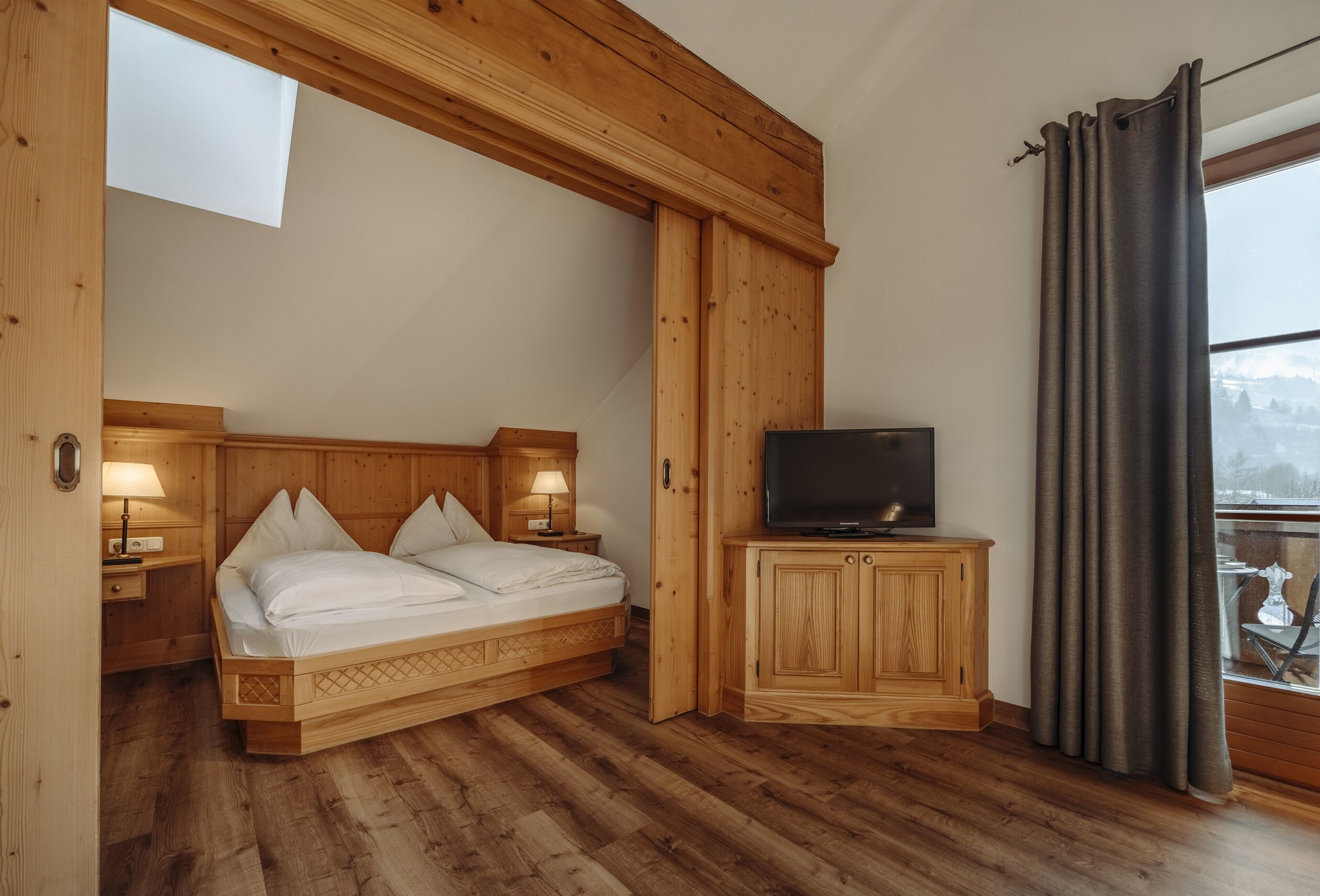Dormitorio con estructura de madera, cama, lámparas de noche, un televisor sobre un mueble de madera y una ventana con cortinas.