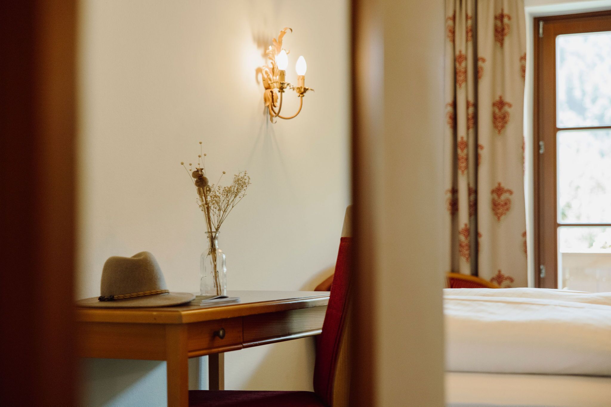 Una habitación con una cama, un escritorio con un sombrero y un jarrón, y una lámpara fijada a la pared.