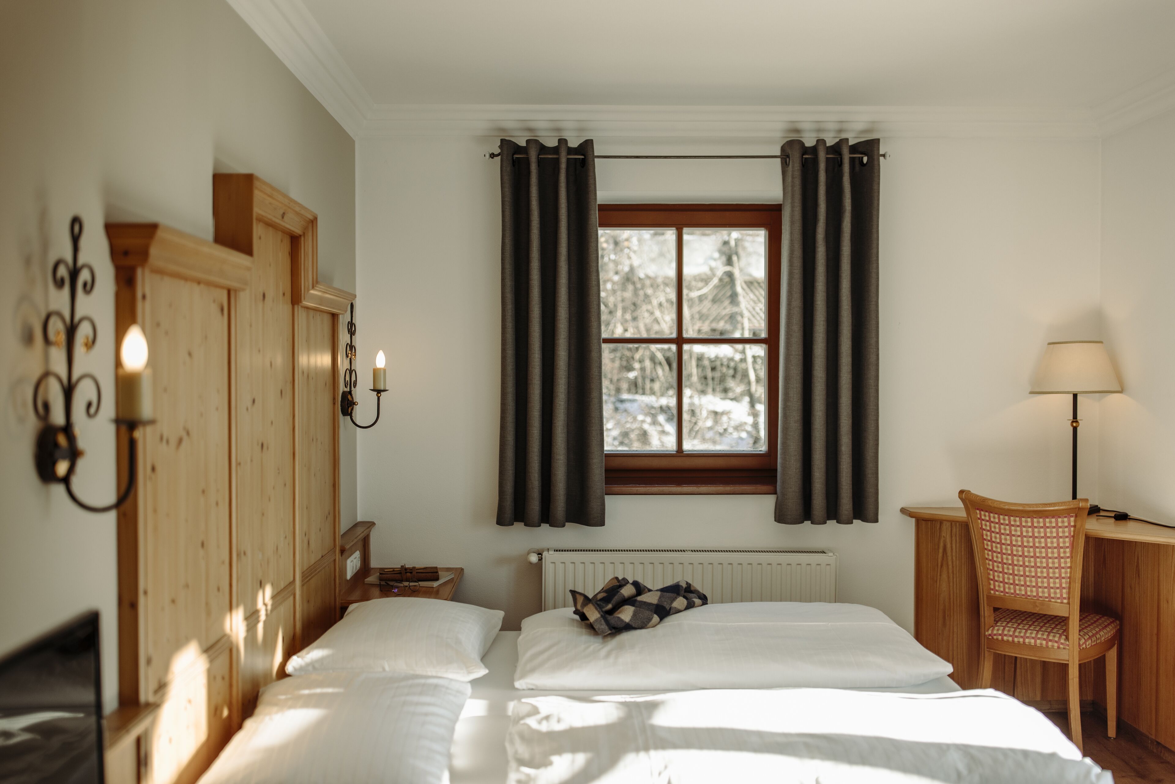 A bedroom with two beds, a window with curtains, a radiator, a desk with a chair, and wall sconces.