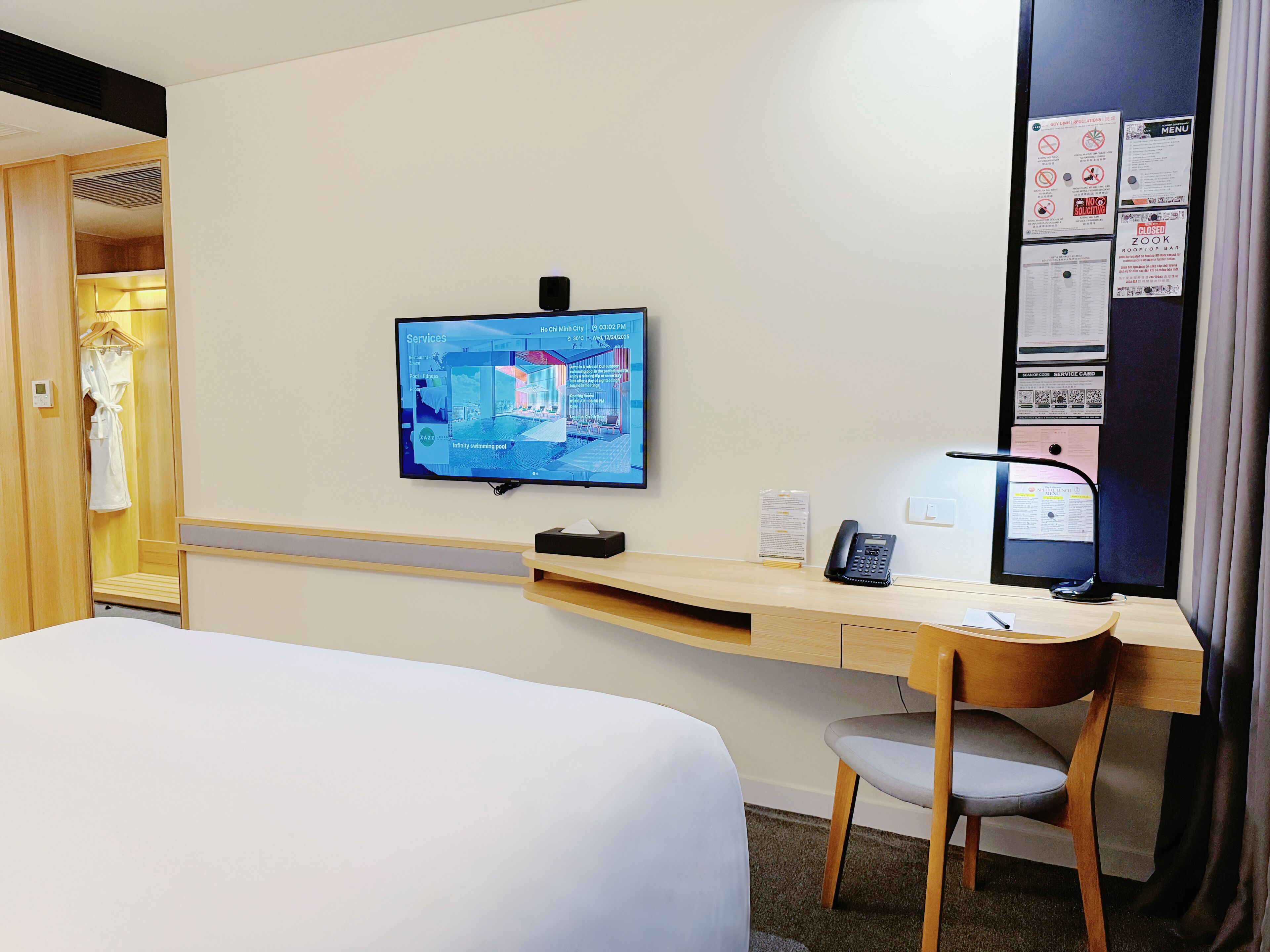 A hotel room with a bed, desk, chair, television, and a bulletin board with notices.
