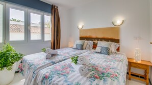A bedroom with two beds, a large window, a headboard with a painting, and a bedside table with a lamp.