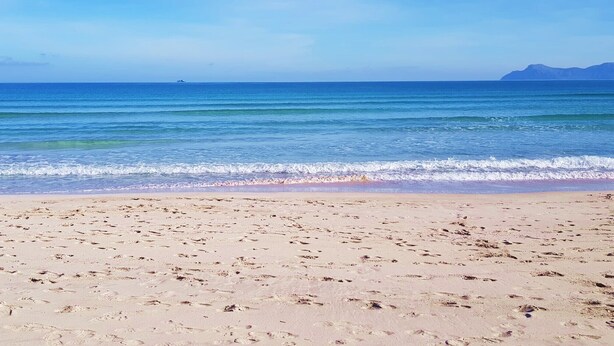 A beach with footprints leading to the water, a clear sky, and distant mountains.