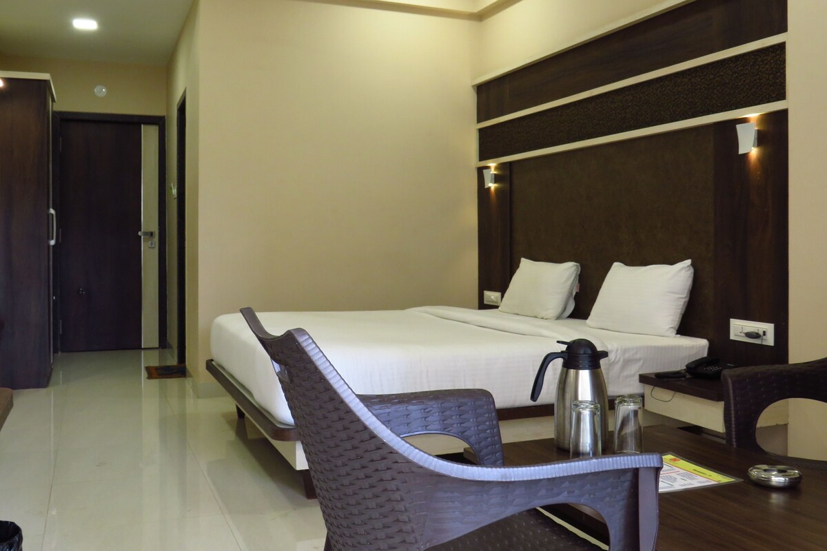 A hotel room with a bed, a chair, a table with a kettle, and a bedside table with a phone.