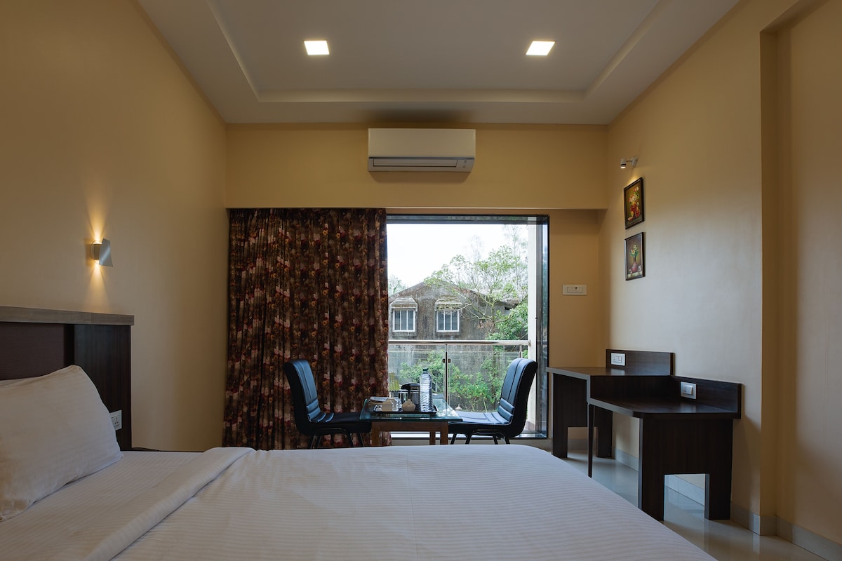 A hotel room with a bed, a desk, two chairs, a window with curtains, and a view of a building outside.
