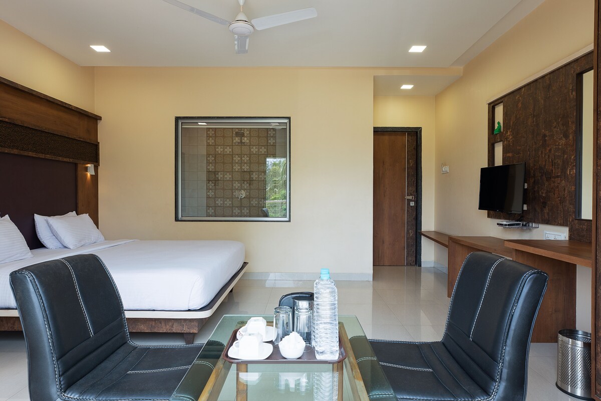 A hotel room with a bed, a desk with a TV, two chairs, a glass table with water bottles, and a ceiling fan.