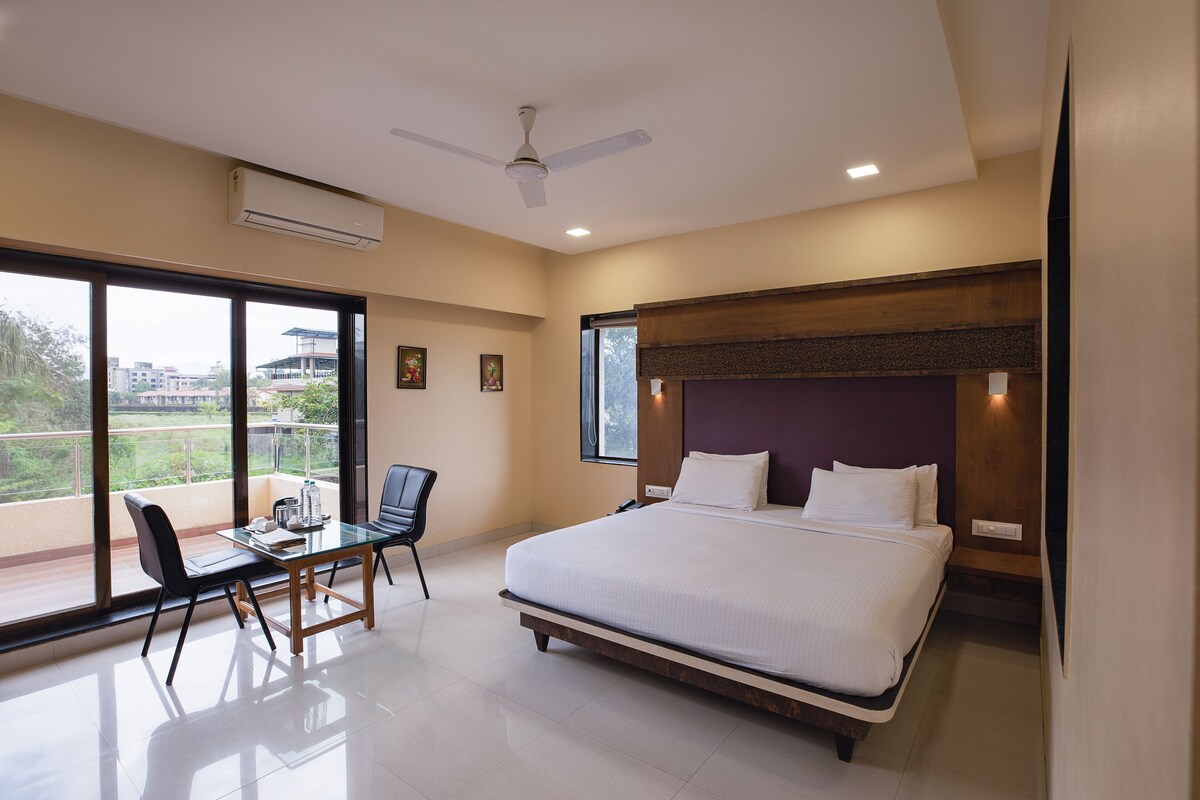 A hotel room with a bed, a desk with a chair, a balcony with a view, and a ceiling fan.