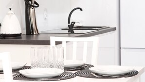 A modern kitchen with a white countertop, a black faucet, and a white chair. There are clear glasses and plates on the counter.
