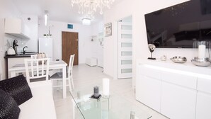 A modern kitchen with white cabinetry, a large flat-screen TV, and a dining area with white chairs and a glass table.