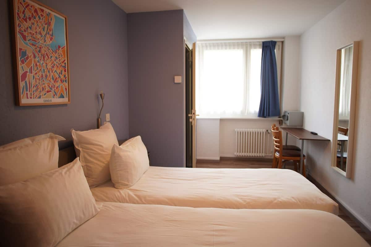 Habitación de hotel con dos camas, un escritorio, una silla, un radiador y una ventana con cortinas.