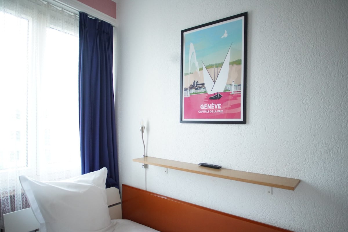 Una habitación de hotel con una cama, un almohadón blanco, una cortina azul, un cuadro de un puente y un estante con un control remoto.
