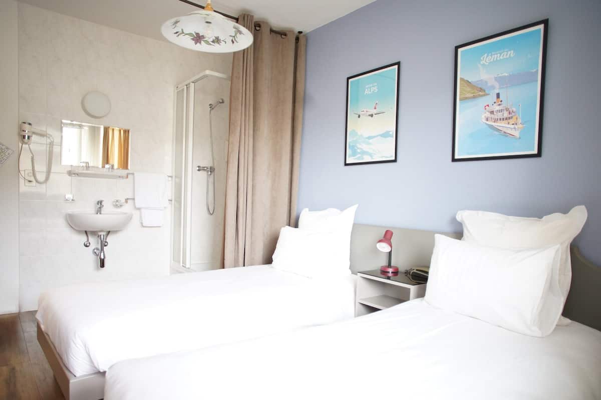 Un cuarto de hotel con una cama grande, una mesita de noche con lámpara, un baño con lavabo y ducha, y dos cuadros enmarcados en la pared.