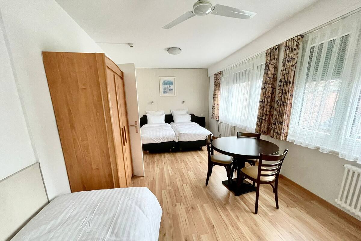Una habitación con dos camas, un armario de madera, un ventilador de techo, una mesa de comedor con sillas y una ventana con cortinas.