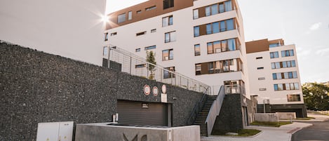 A modern building with multiple floors, a staircase leading to the entrance, and a paved area with a metal fence.