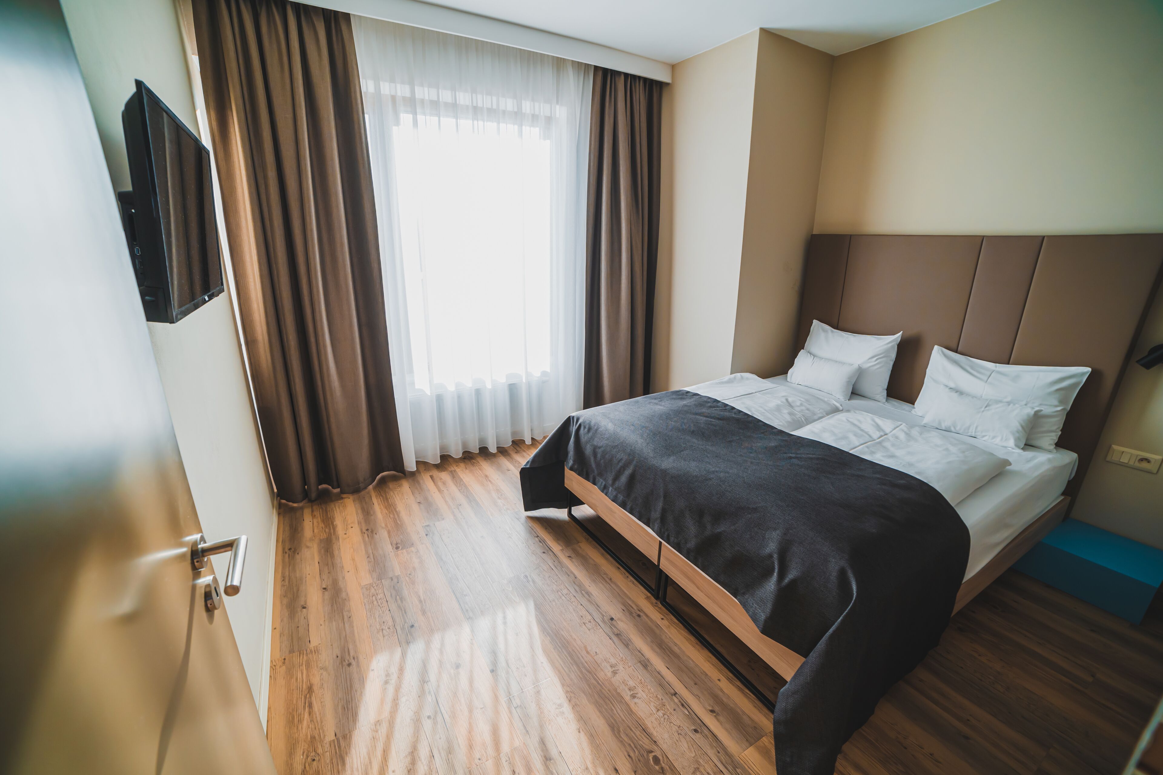 A hotel room with a wooden floor, a bed with white linens, a dark headboard, and a window with curtains.