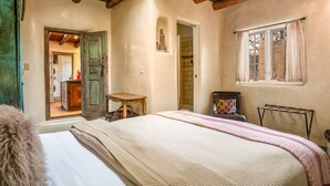 A bedroom with a wooden ceiling, a bed with a quilt, a small table, a window with curtains, and a door leading to another room. - Authentic Santa Fe Home-Step into History & Charm (Santa Fe)