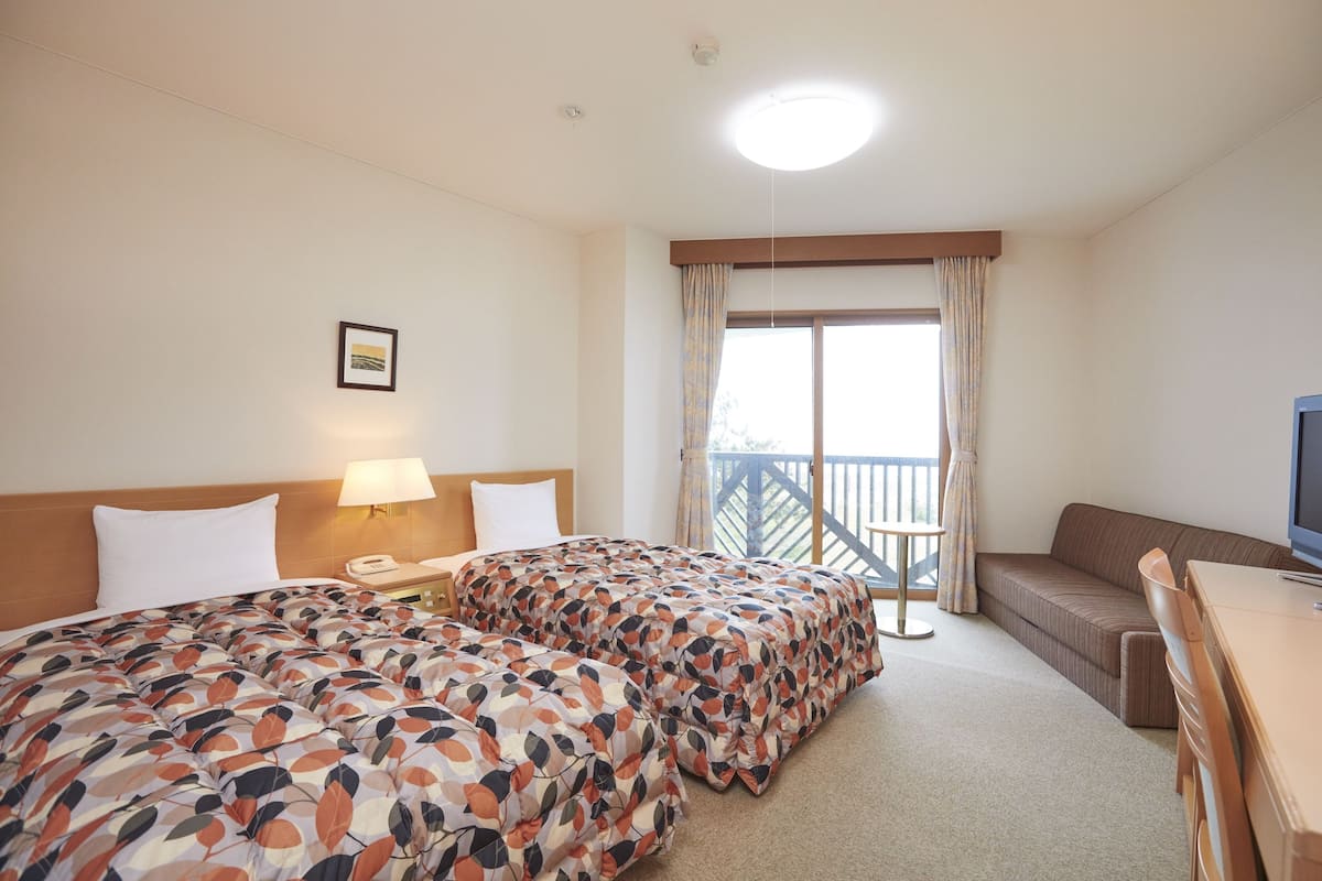 A hotel room with two beds, a sofa, a TV, a balcony with curtains, and a small table.